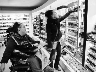 Eine Frau im Elektrorollstuhl wird von ihrer Assistentin dabei unterstützt, Kosmetikprodukte einzukaufen